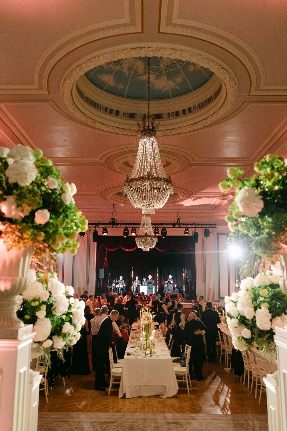 A candlelit wedding reception under a chandelier, with a band playing and long tables set between tall white florals