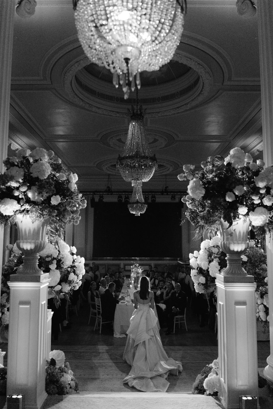 A bride walking into a candlelit ballroom beneath crystal chandeliers